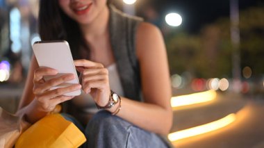 Gülümseyen modern kadın cep telefonuna mesaj yazarken arka planda bulanık ışıklarla City Caddesi 'nde oturuyor..