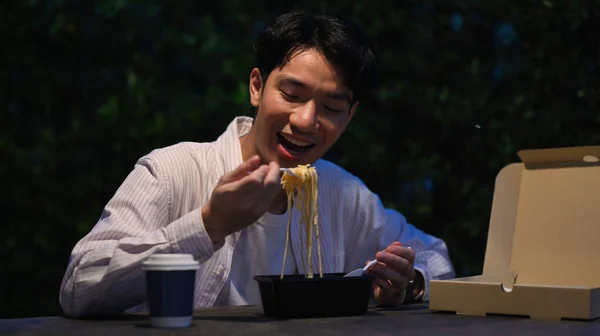Happy young asian man eating take away noodles at a table at night market. Nightlife concept.