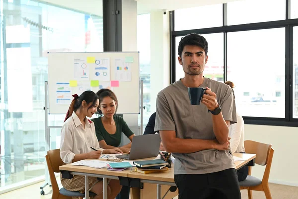 Handsome man team leader sitting in modern start up office with group of colleagues in the background.