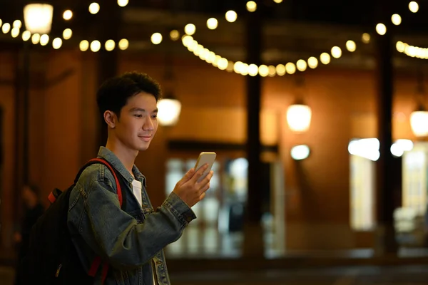 Uśmiechnięty Azjata Sprawdza Swój Telefon Komórkowy Stojąc Miejskiej Alei Rozmytymi — Zdjęcie stockowe