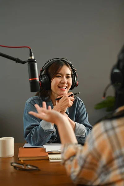 Friendly female radio host looking and talking with her guest while making audio podcast in studio.