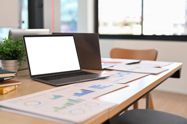 Laptop computer, financial graph data documents on wooden table in bright office.