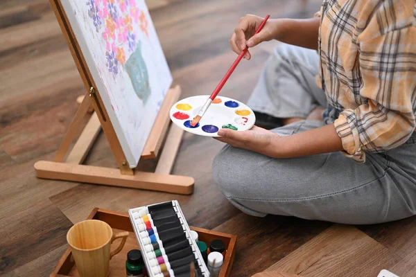 Cropped image of young female artist holding palette and painting picture on canvas with oil paints in home studio.