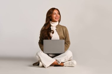 Moda kıyafetleri içinde güzel bir kadın beyaz stüdyoda dizüstü bilgisayarla oturuyor. İnsanlar ve teknoloji kavramı.
