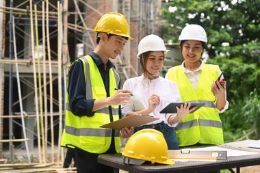 Mühendis ekibi ve yatırımcı dijital tablet kullanarak, arka plandaki tamamlanmamış konut binasının önündeki plan detaylarını tartışıyor.