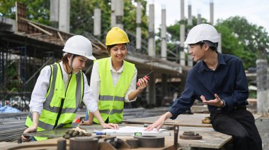 Güvenlik kaskı takan bir grup mühendis inşaat alanında çalışıyor, planı birlikte kontrol ediyorlar.. 