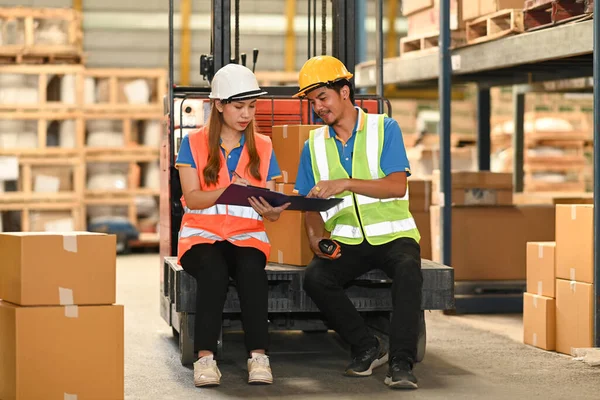 Two warehouse workers wearing safety hard hat and vest sitting on forklift truck in retail warehouse checking newly arrived goods.