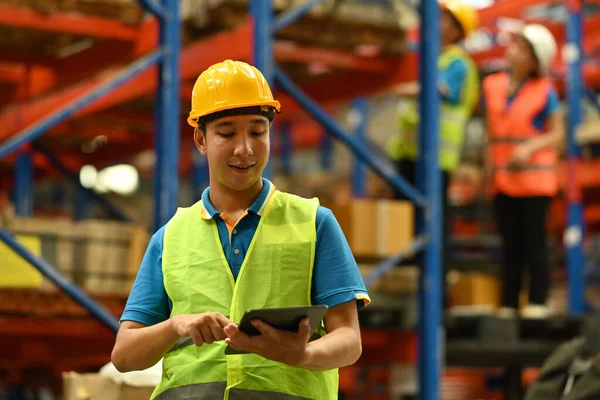 Asian male manager in hardhat and reflective jacket using digital tablet, checking inventory production stock control in warehouse.