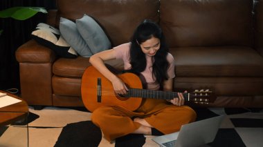 Mutlu genç bir kadın akustik gitar çalıyor, sıcak oturma odasında hafta sonunun tadını çıkarıyor..