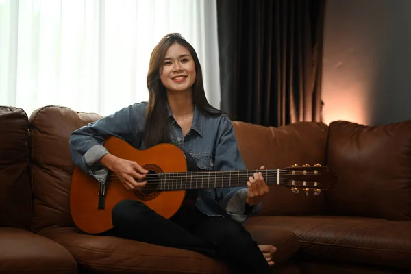 소파에 앉아서 어쿠스틱 기타를 여가를 집에서 보내는 여자에게 미소를 — 스톡 사진