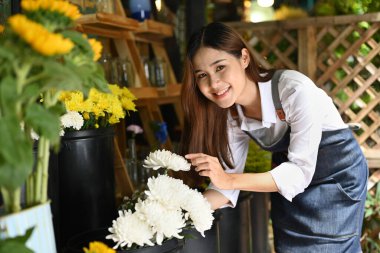 Başarılı küçük işletme girişimcisi önlüklü, kır çiçekçisinde elinde çiçeklerle dikiliyor..