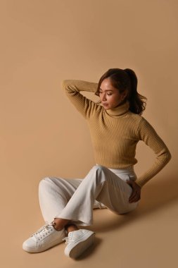 Güzel, genç bir kadın bej arka planda poz veren, toprak renginde kalın kıyafetler giyiyor. Sonbahar ve Kış kavramı.