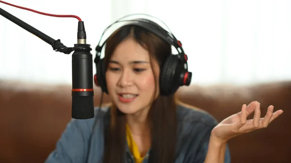 Asian woman radio host wearing headphone and talking through microphone recording podcast in small home studio.