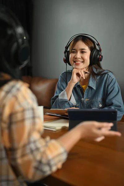 Shot of smiling young female radio host listening to interesting conversation with guest during recording podcast in home studio.