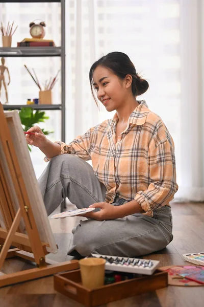 Satisfied woman sitting on floor in home studio and painting picture on canvas. Leisure activity, creative hobby and art concept.