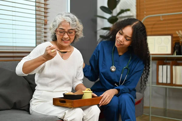 健康的な栄養の朝食を持っている高齢者の女性の写真午前中にソファの上にプロの介護者と食事 — ストック写真