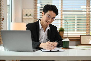 Mutlu genç işadamı dizüstü bilgisayar kullanıyor ve not defterine önemli bilgiler yüklüyor..