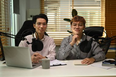 Yoğunlaştırıcı mikrofonun önünde oturup kameraya gülümseyen iki mutlu podcaster resmi. Radyo, podcast ve teknoloji konsepti.