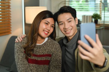 Gülümseyen genç bir çift selfie çekiyor, akıllı telefondan video çekiyor, evdeki kanepede oturuyor..