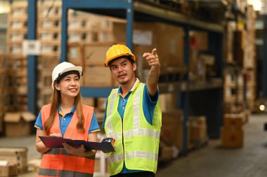 Perakende deposunda uzun raflar arasında duran kasklı ve yansıtıcı ceketli iki depo çalışanı..
