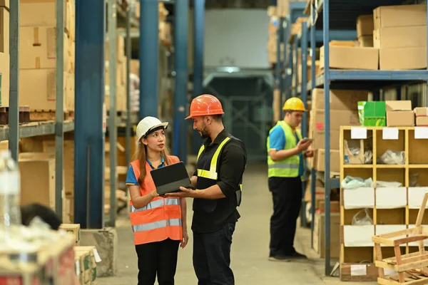 Full length of two warehouse workers are standing between retail warehouse full of shelves and checking order details on a tablet.