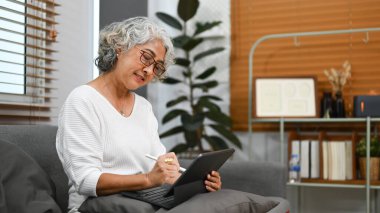 Pozitif orta yaşlı kadın dijital tablette kablosuz internete bakıyor, evdeki kanepede dinleniyor..