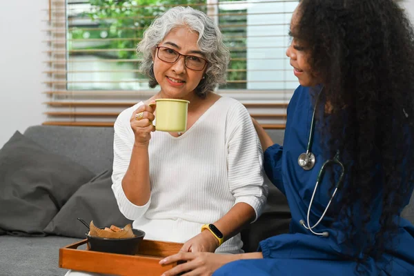 笑顔の高齢者の女性は 午前中にソファの上に有用な介護者と健康的な栄養朝食の食事を持っている — ストック写真