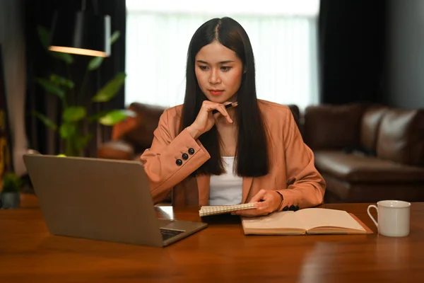 Thoughtful businesswoman reading online information on laptop computer while working remote from home.