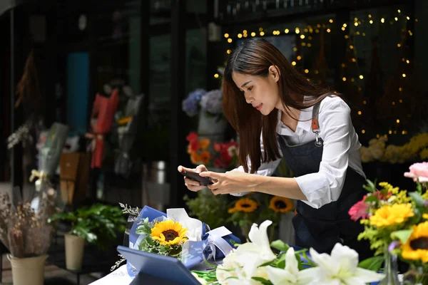 Asian woman small business entrepreneur using smart phone taking picture of bouquet of fresh flowers for client order.