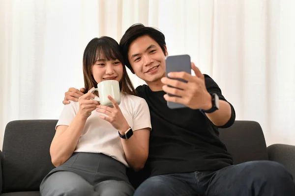 幸せな若いですカップル作る自画撮りとともにスマートフォンでリラックスしながらソファで明るいリビングルーム — ストック写真