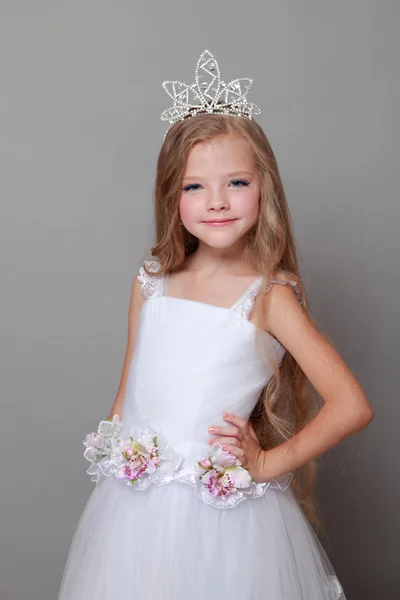 Linda niña usando una corona y un vestido blanco lindo sonriendo y posando para la cámara sobre un fondo gris —  Fotos de Stock