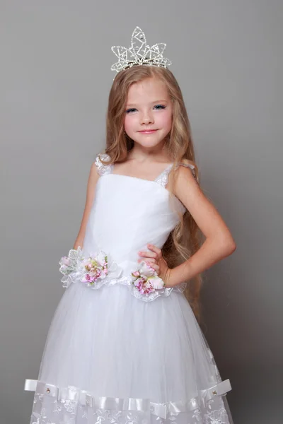 Mała dziewczynka kaukaski z piękne długie włosy w koronie i białą sukienkę z ładny uśmiech, pozowanie na kamery na szary tło uroda i moda — Zdjęcie stockowe