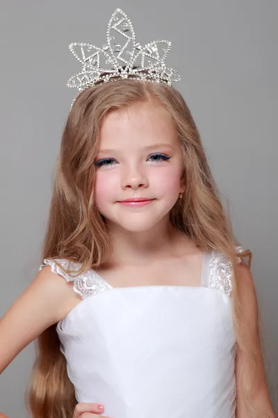 Niña caucásica con hermoso pelo largo en la corona y un vestido blanco con una linda sonrisa posando en la cámara sobre un fondo gris en la belleza y la moda —  Fotos de Stock