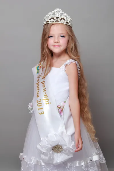 Carino bambina indossa una corona e un vestito bianco carino sorridente e in posa per la fotocamera su uno sfondo grigio — Foto Stock