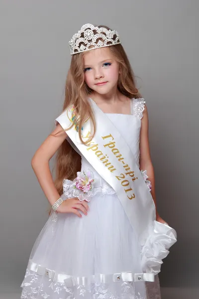 Happy little girl with long hair wearing a crown and a white dress with a ribbon and the words "Grand Prix of Ukraine 2013" on Beauty and Fashion — Stock Photo, Image