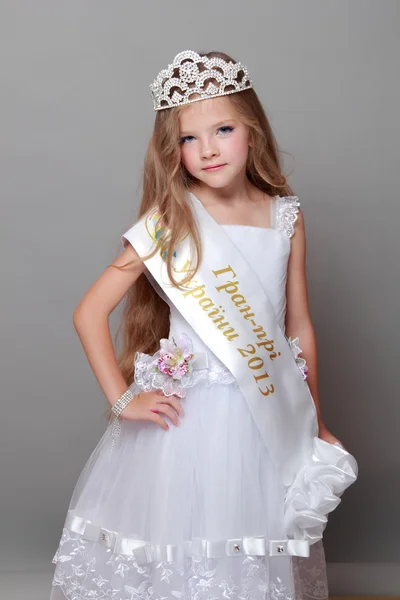 Happy little girl with long hair wearing a crown and a white dress with a ribbon and the words "Grand Prix of Ukraine 2013" on Beauty and Fashion — Stock Photo, Image