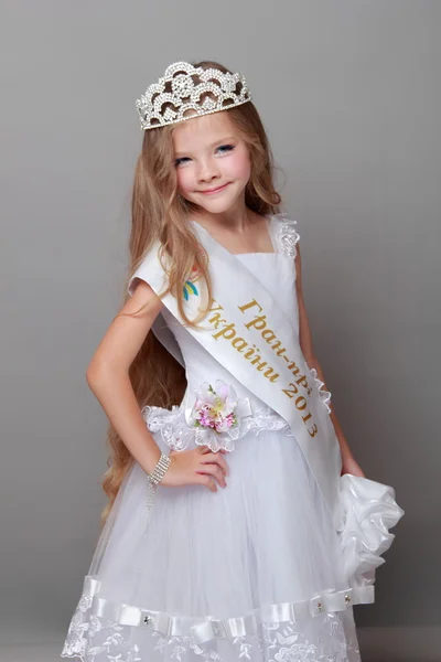 Linda niña usando una corona y un vestido blanco lindo sonriendo y posando para la cámara sobre un fondo gris —  Fotos de Stock