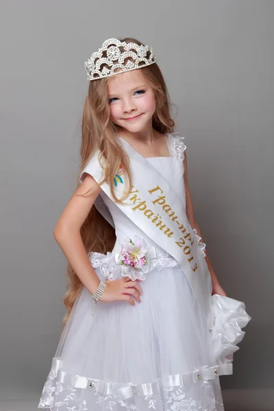 Cute little girl wearing a crown and a white dress cute smiling and posing for the camera on a gray background — Stock Photo, Image