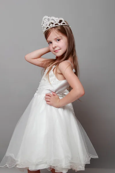 Mała dziewczynka kaukaski z piękne włosy w koronie i białą sukienkę z ładny uśmiech, pozowanie na kamery na szarym tle — Zdjęcie stockowe