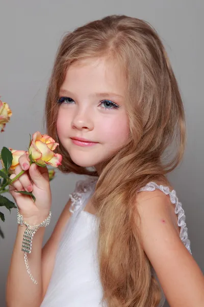 Petite fille souriante européenne dans une robe blanche aux cheveux longs tient une rose fraîche saine avec des feuilles vertes sur la beauté et la mode — Photo