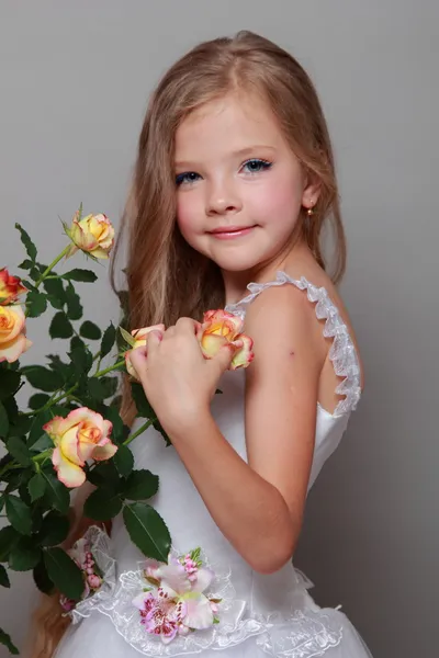 Petite fille souriante européenne dans une robe blanche aux cheveux longs tient une rose fraîche saine avec des feuilles vertes sur la beauté et la mode — Photo