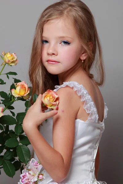 Europäisches lächelndes kleines Mädchen in einem weißen Kleid mit langen Haaren hält gesunde frische Rosen mit grünen Blättern über Schönheit und Mode — Stockfoto
