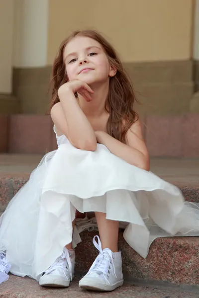 Elegante schattig klein meisje in een witte jurk met mooie haren in sneakers op schoonheid en mode — Stockfoto