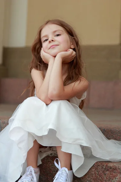 Güzel beyaz topu cüppe ve spor ayakkabı için belgili tanımlık açık havada merdivenlerde oturan neşeli küçük kız gülümseyerek — Stok fotoğraf