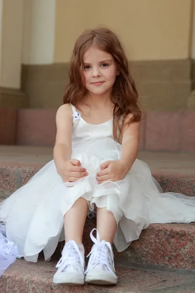 Smiling cheerful little girl in a beautiful white ball gown and sneakers sitting on the stairs to the outdoors — Stock Photo, Image