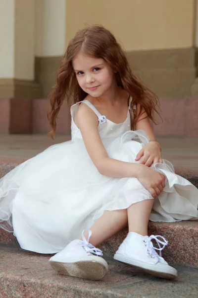 Lächelndes fröhliches kleines Mädchen in einem wunderschönen weißen Ballkleid und Turnschuhen, das auf der Treppe nach draußen sitzt — Stockfoto