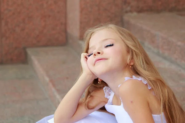 Portrait of cute smiling little girl with long hair in a beautiful dress is sitting on the steps outdoors — Stock Photo, Image