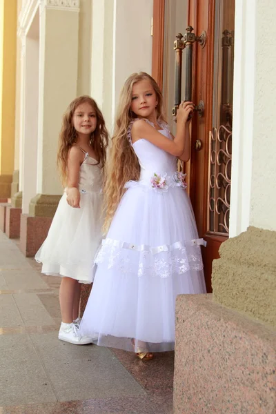Dvě krásné holčičky v dlouhé bílé šaty stojí poblíž zrcadlové dveře budovy venku — Stock fotografie