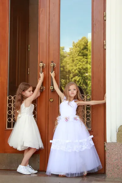 Duas meninas encantadoras em vestidos brancos longos ficam perto das portas espelhadas do edifício ao ar livre — Fotografia de Stock