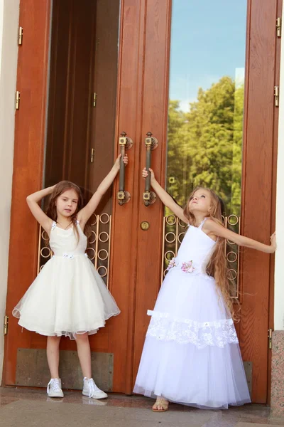 Dos encantadoras niñas con largos vestidos blancos están cerca de las puertas espejadas del edificio al aire libre — Foto de Stock